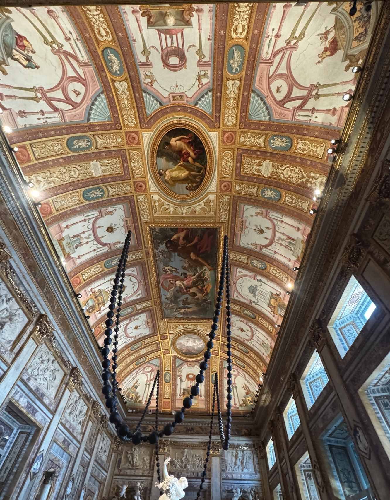 Ceiling view inside Galleria Borghese in Rome, showing ornate frescoes, gold architectural details, and a contemporary Wangechi Mutu installation suspended above classical sculptures. Photo by Barbara Rozgonyi